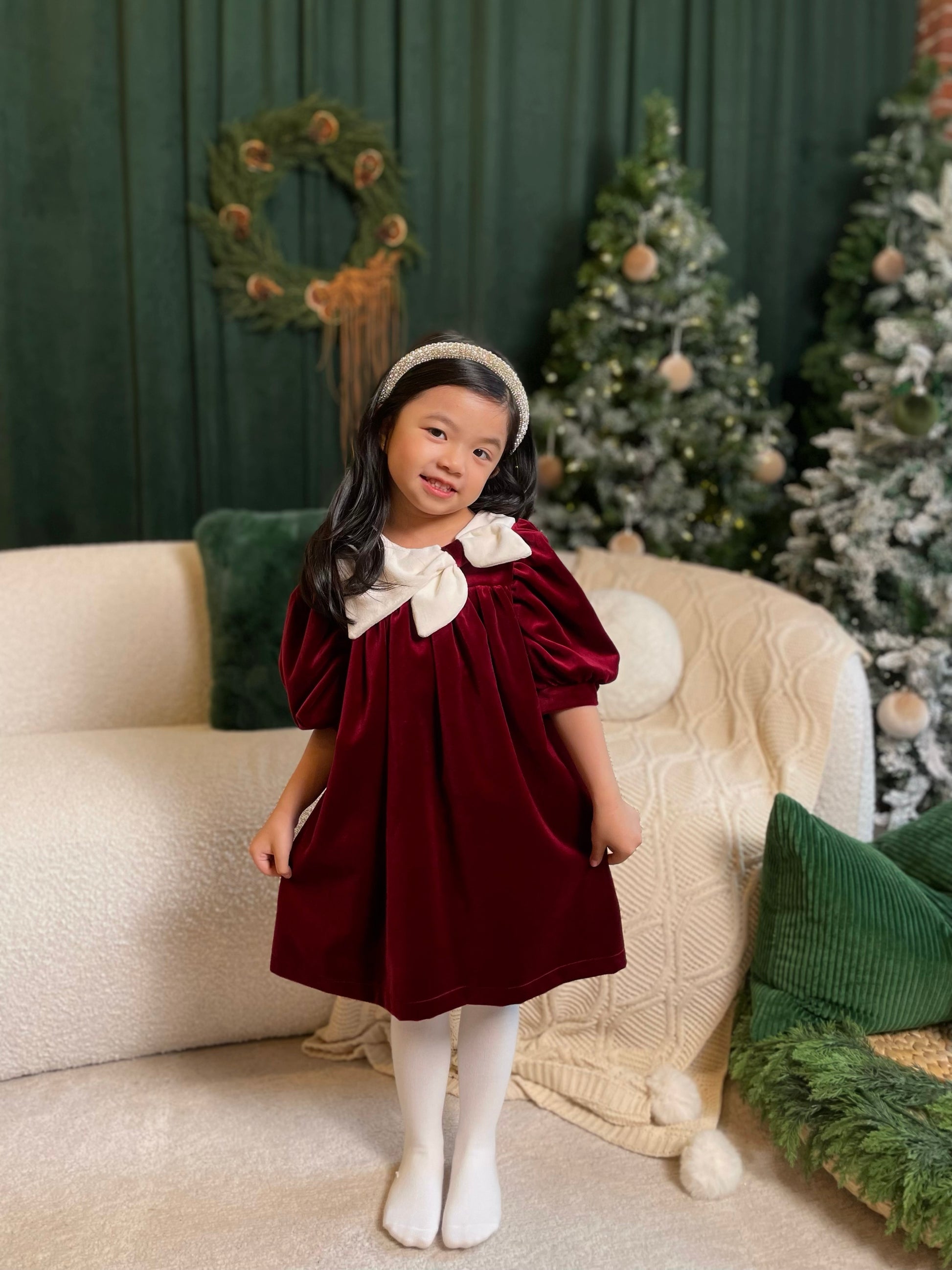 Young girl in a burgundy dress with white collar standing in a festive room with Christmas trees and decorations.