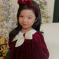 Young girl in a burgundy dress with a white bow and red headband, standing in front of a decorated Christmas tree.