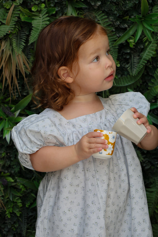 Child in a light dress holding a cup and saucer against a green leafy background