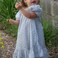 Young girl in a light blue floral dress standing outdoors with greenery and a stone wall in the background.