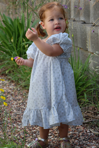 Young girl in a light blue floral dress standing outdoors with greenery and a stone wall in the background.