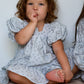 Child in a light blue floral dress sitting on a beige cushion against a white wall.