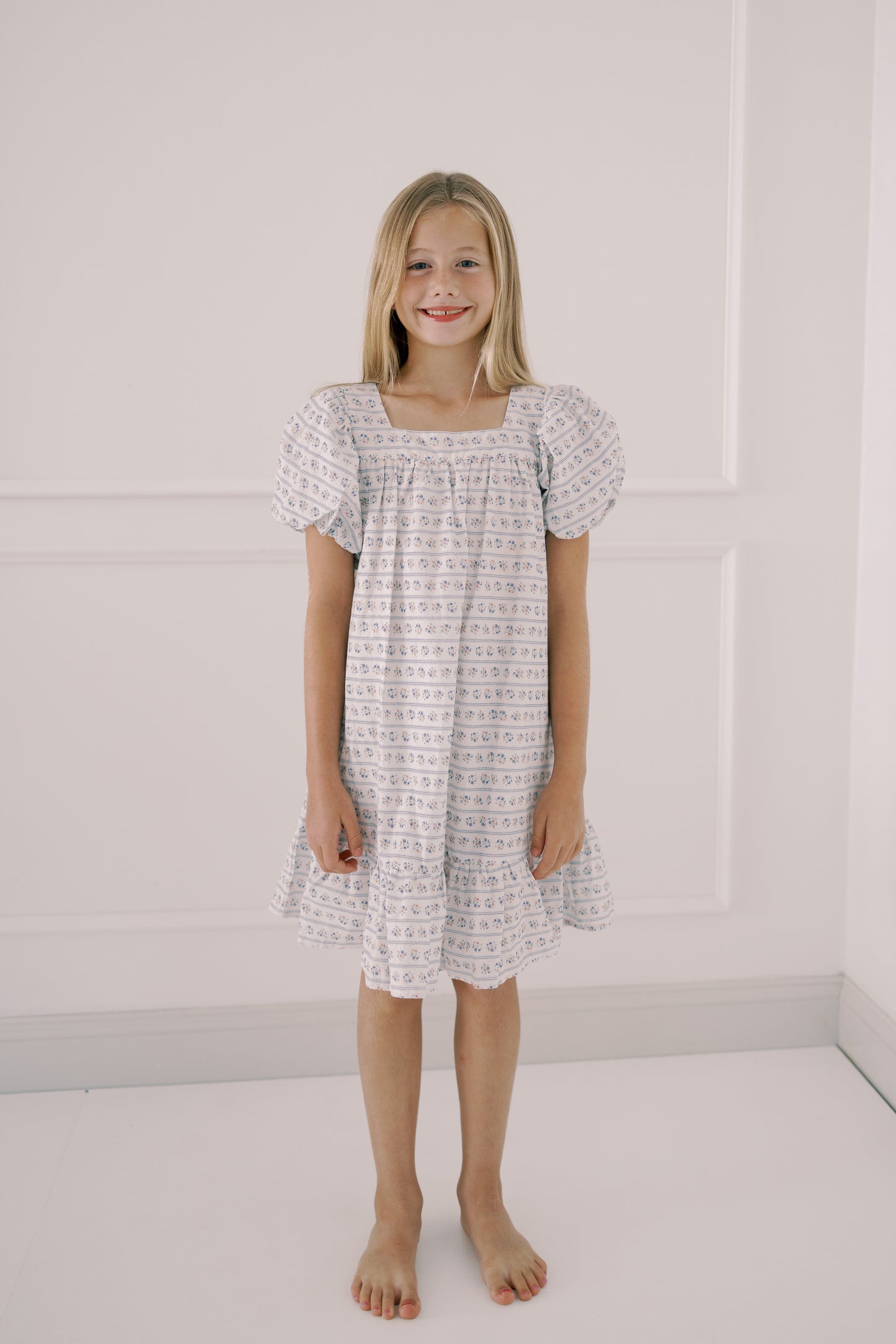 Young girl wearing a patterned dress standing against a white wall.