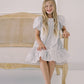 Young girl in a white patterned dress sitting on a wooden chair against a white background