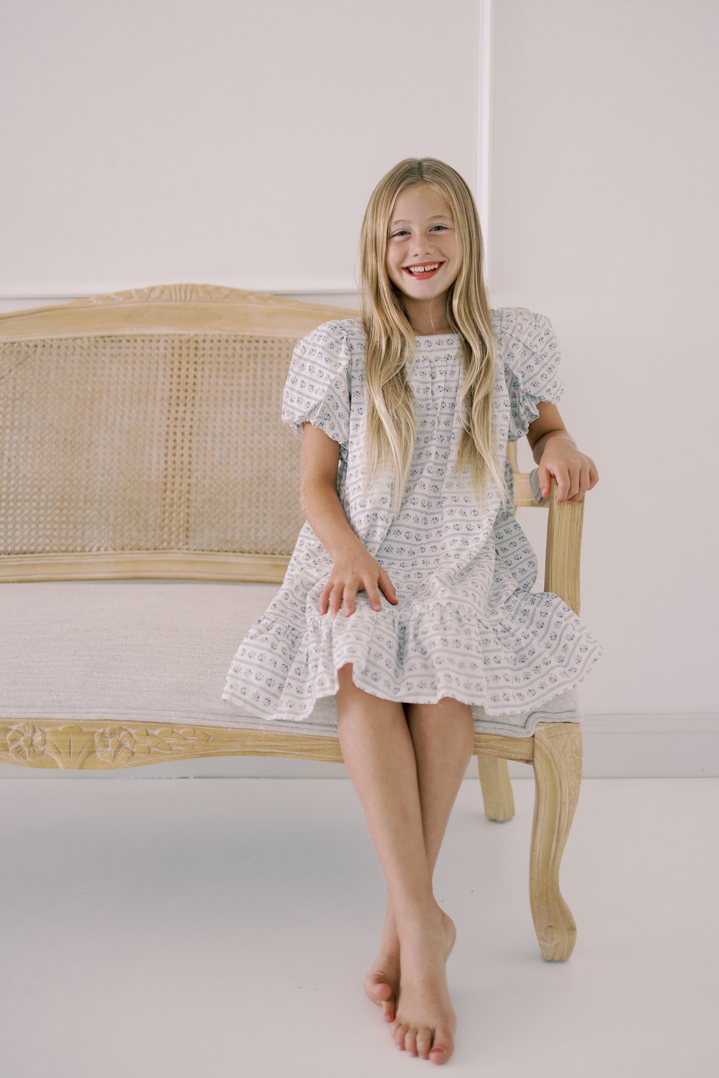 Young girl in a white patterned dress sitting on a wooden chair against a white background