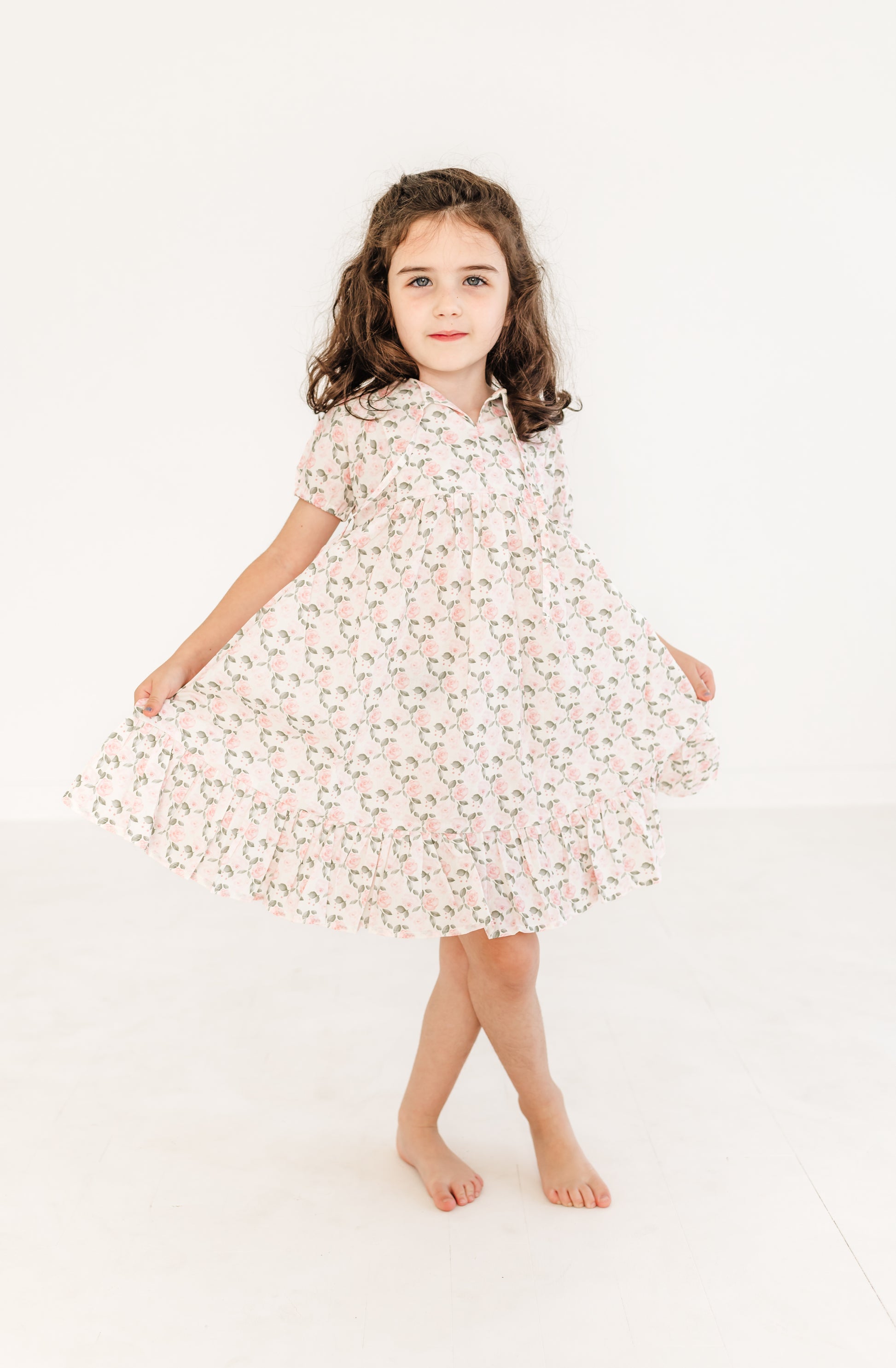 A young girl standing against a white background, wearing a beige floral print dress with short sleeves and a knee-length skirt.